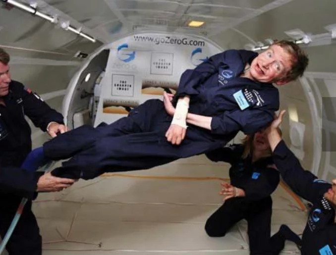 A group of people in blue jumpsuits assisting a man in a similar outfit as he experiences weightlessness in a zero-gravity simulator. The interior is designed for space training with equipment visible.