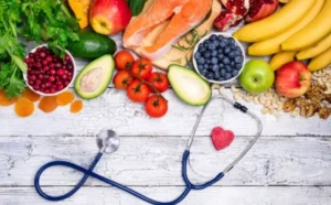 An assortment of fresh fruits, vegetables, and salmon arranged around a stethoscope and a small red heart shape, symbolizing health and nutrition on a wooden surface.