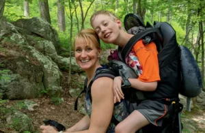A woman smiling while carrying a young boy in a backpack on a wooded hiking trail. The background features green trees and rocky terrain.