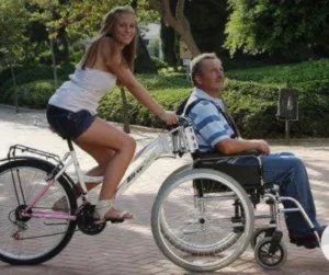 A young woman rides a bicycle while leaning over a man in a wheelchair. They are outdoors on a brick path surrounded by greenery.