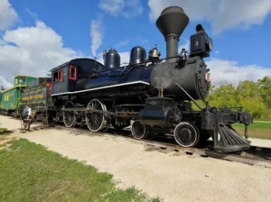 A vintage steam locomotive, black with red accents, parked on a gravel track. In the background, another train car is visible. The scene is set against a blue sky with fluffy clouds, emphasizing the historical aspect of railway transport.