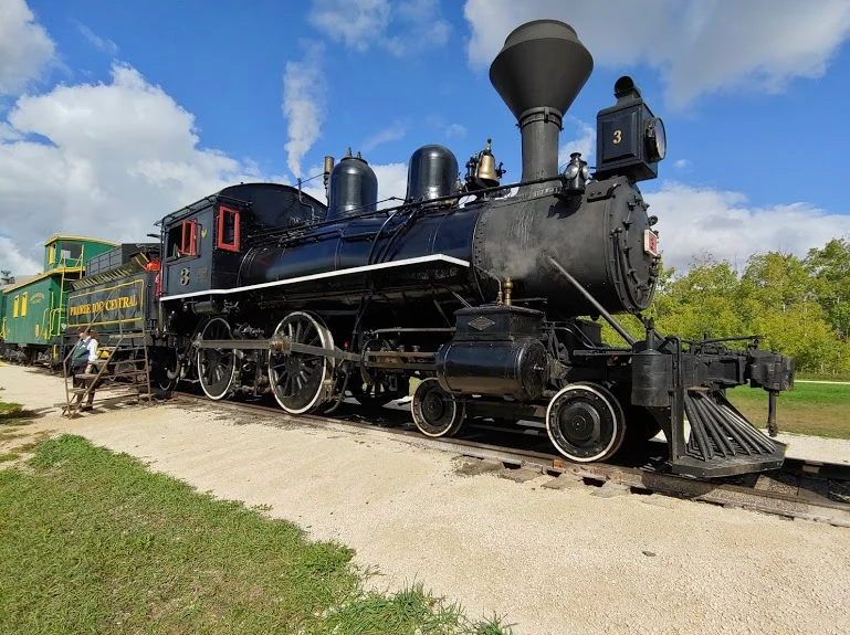 A vintage steam locomotive, black with red accents, parked on a gravel track. In the background, another train car is visible. The scene is set against a blue sky with fluffy clouds, emphasizing the historical aspect of railway transport.