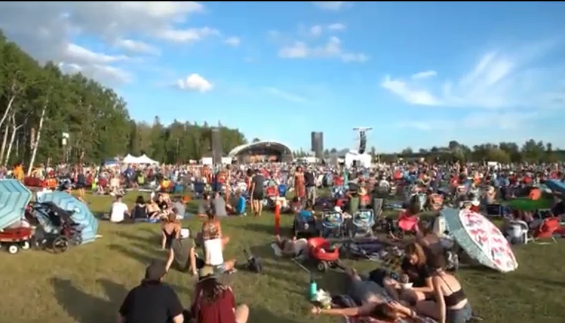 A large crowd of festival attendees enjoying an outdoor music event at the Winnipeg Folk Festival, with tents, stage structures, and people seated on the grass under a blue sky.