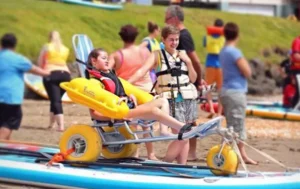 A young girl seated in a beach wheelchair with large yellow wheels, assisted by an adult in a life jacket. Other people are preparing for water activities in the background on a sandy beach.