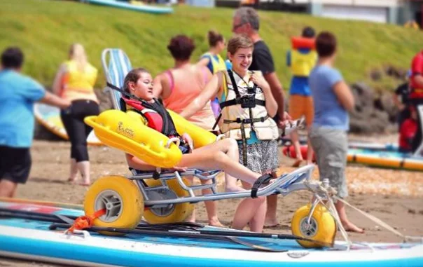 A young girl seated in a beach wheelchair with large yellow wheels, assisted by an adult in a life jacket. Other people are preparing for water activities in the background on a sandy beach.