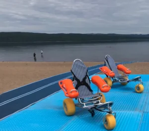 Two wheeled water chairs with flotation devices on a blue mat at a beach. In the background, two people are walking along the shoreline, with calm water and mountains in the distance under a cloudy sky.