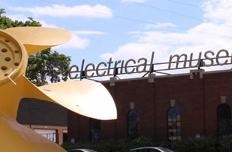 A large yellow structure resembling a turbine is in the foreground, with the text 'electrical museum' visible on a building in the background under a blue sky.