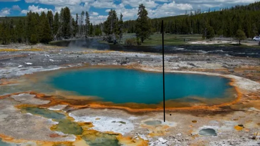 A vibrant hot spring with turquoise water surrounded by orange mineral deposits and barren ground. In the background, green trees and a distant forested area are visible under a partly cloudy sky.