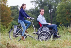 A couple rides a bicycle with a wheelchair attached, promoting accessibility and shared outdoor experiences.