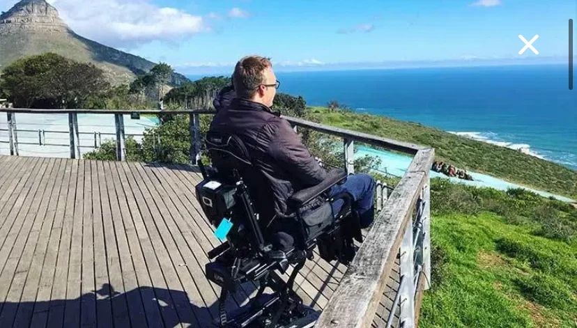 A person in a wheelchair sits on a wooden deck overlooking a scenic view of the ocean and hills under a blue sky.