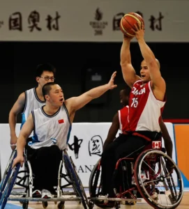 Athletes in wheelchairs playing basketball during a competitive match. A player in a red jersey with 'Canada' written on it holds the ball while opponents attempt to block.