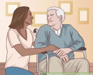 A woman seated beside an elderly man in a wheelchair, both smiling and engaged in conversation in a well-lit room with a simple background.