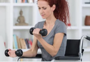 A woman sitting in a wheelchair is exercising with dumbbells in a bright room. She has curly red hair and is focused on her workout, wearing a gray t-shirt.