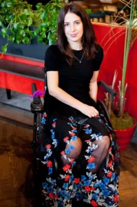 A woman with long brown hair sitting in a wheelchair, wearing a black top and a floral skirt, posing in a vibrant indoor setting with plants and red accents.