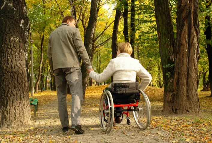 A man holding hands with a woman in a wheelchair while walking on a path through a park with autumn foliage.