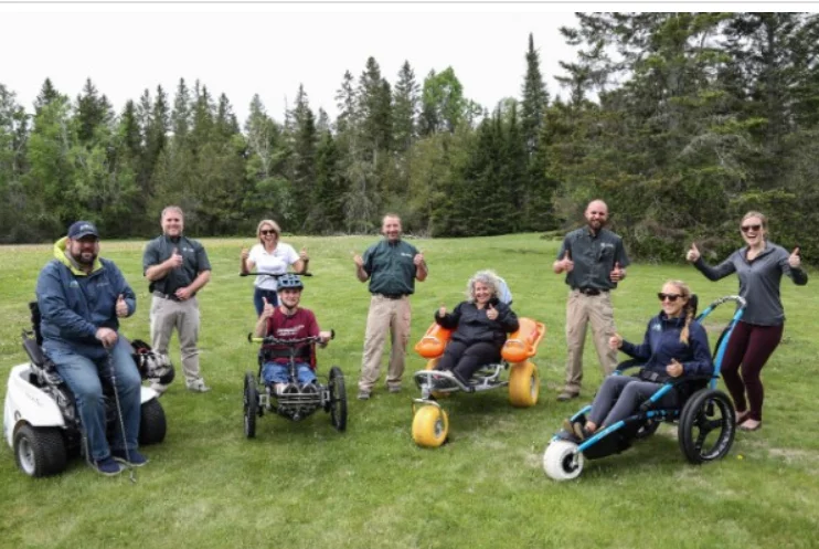 A group of eight individuals, both adults and a child, gathered in a grassy area. Some are on adaptive mobility devices, while others stand behind, all smiling and giving thumbs up. Pine trees are visible in the background.