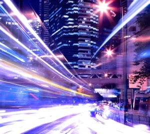 A cityscape at night featuring tall buildings illuminated by lights, with streaks of light from moving vehicles creating a dynamic effect on a busy street.