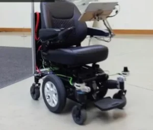 An electric wheelchair with a black seat and armrests, showing visible wiring and a control panel mounted on the back. It is positioned on a light-colored floor in an indoor setting.