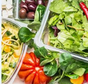 A variety of fresh ingredients including a bowl of pasta with vegetables, a mixed salad, olives, and a ripe tomato arranged on a white surface.
