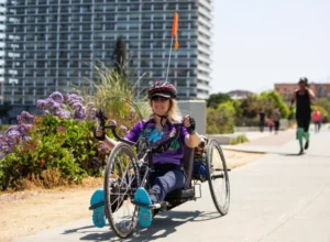 A woman riding a handcycle on a paved path lined with purple flowers, wearing sunglasses and a helmet, with a flag attached to her bike. In the background, there are modern buildings and other people walking.