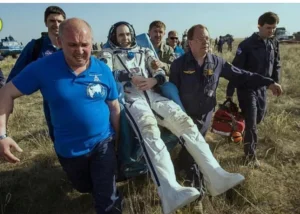 A group of people assisting a space astronaut in a space suit, who is being carried on a stretcher after landing. The scene takes place in a grassy area, with team members in uniform visible around them.