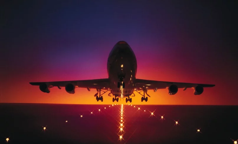 A silhouette of an airplane landing against a vibrant sunset with shades of purple and orange. Lights are visible on the runway, creating a guiding path for the aircraft.
