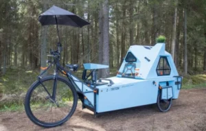 A bicycle with a light blue cargo trailer attached, featuring a compact cabin-like structure with a small umbrella and greenery on top, situated on a dirt path in a wooded area.