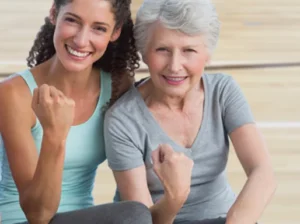 A young woman and an older woman sitting together on a gym floor, both smiling and raising their fists in a gesture of celebration or encouragement. The background shows a wooden gymnasium floor.