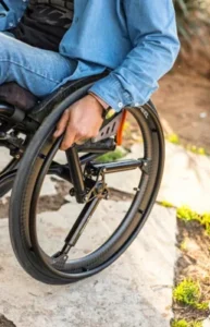 A close-up view of a person's hand gripping the wheel of a manual wheelchair, with the person wearing a denim shirt and blue jeans. The wheelchair is positioned on a stone surface near grass.