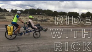 A person in a yellow helmet rides a tandem bicycle while pulling a passenger wearing a white shirt and gray pants. The scene takes place on a rural road surrounded by sparse vegetation and cloudy skies.