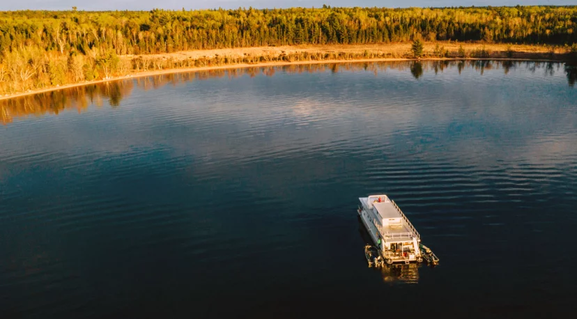 Aerial view of a boat floating on a calm lake surrounded by lush green trees, reflecting the blue sky and the shoreline.