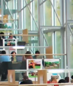 An indoor workspace with multiple levels, featuring people working at desks with computers. Large windows provide natural light, and colorful posters are displayed on nearby walls.
