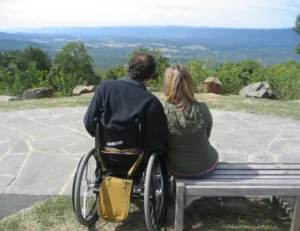 A man in a wheelchair and a woman sitting next to him on a bench, looking out at a scenic landscape with mountains and greenery in the distance. This image highlights the challenges faced by caregiver spouses.