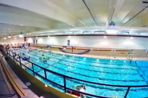 A panoramic view of an indoor swimming pool with multiple lanes, where people are swimming and floating on colorful pool toys. Benches line the side of the pool.