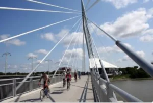 Cyclists riding along a modern pedestrian bridge with cable stays, crossing over a river on a sunny day. The scene includes a blue sky with clouds and lush greenery in the background, part of the Red and Assiniboine Riverwalk Promenades.