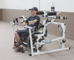 A person in a wheelchair using an adaptive exercise machine, focusing on upper body strength training in a spacious gym environment. The machine includes various attachments for targeted workouts.