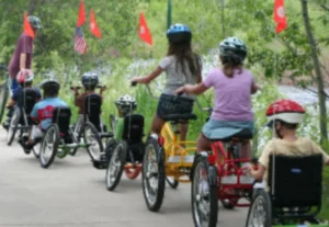 A group of children riding adaptive tricycles along a paved path, surrounded by trees. Some are wearing helmets and colorful shirts. Red flags are visible along the path.