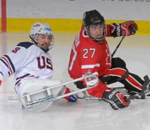 Two sled hockey players competing on the ice. One player wears a white jersey with 'USA' and the other wears a red jersey with '27' highlighted. They appear to be maneuvering during the match.