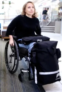 A woman in a wheelchair, dressed in black, rolls through a shopping center hallway. She has a large black backpack attached to her chair, and the environment features bright indoor lighting and people in the background.