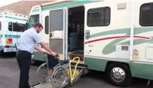 A man in a uniform is operating a wheelchair lift on the side of a recreational vehicle, helping to load a wheelchair onto the vehicle. The scene shows the vehicle parked in a lot.