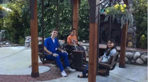 Two young adults sitting on swings in a garden, one in a wheelchair holding a toy, the other in blue scrubs smiling. A third person sits to the right, wearing a sweater, with greenery and flowers in the background.