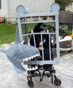 A person in a wheelchair dressed in a shark costume, with a gray cage design around them. The costume features a large shark head with sharp teeth at the front, positioned on a paved outdoor area.