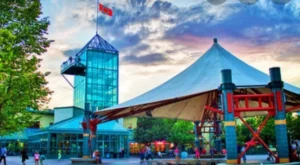 A modern building with a glass structure and a Canadian flag, beside a large tent-like structure supported by red poles. People are gathered in the area, with trees and a cloudy sky in the background.