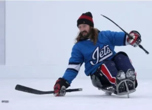 A man in a Winnipeg Jets jersey sits on a sled on ice while holding a hockey stick. He wears a black beanie and smiles as he skates, demonstrating accessible outdoor hockey in the Manitoba community.
