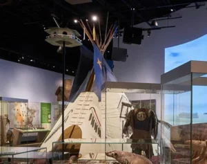 An indoor museum display featuring a traditional teepee with a blue flag on top, surrounded by glass cases containing various artifacts and a mural of a natural landscape in the background.