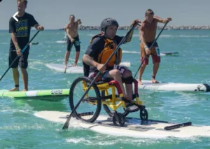 A person in a wheelchair on a paddleboard, using a paddle to navigate through water. Several other individuals are standing on paddleboards in the background, enjoying the activity on a sunny day.