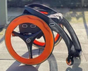 A modern wheelchair featuring large wheels with orange accents, positioned on a patio surface. The design showcases a sleek frame with a black seat and handles.