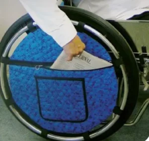 A person reaching into a storage pouch attached to the wheel of a wheelchair, retrieving a newspaper. The wheel features a blue decorative cover with a pocket.