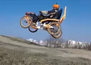 A person sitting in a modified bicycle with an attached seat, performing a jump over a grassy hill against a clear blue sky.