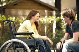 A woman in a wheelchair smiling at a man sitting across from her in a garden setting. Both appear engaged in conversation, with greenery and a structure visible in the background.